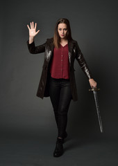 full length portrait of brunette girl wearing long leather coat and boots. standing pose and holding a sword on grey studio background.