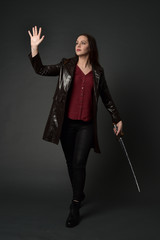 full length portrait of brunette girl wearing long leather coat and boots. standing pose and holding a sword on grey studio background.