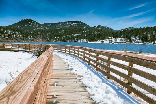 VIEW WOODEN WALK WAY WITH BACKGROUND OF BRIGHT BLUE SKY AND  SNOW ON GROUND OF EVERGREEN LAKE IN LATE WINTER / COLORADO / DENVER