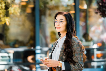 beautiful brunette woman in autumn outfit holding smartphone and looking away on street near cafe