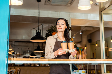 beautiful waitress in apron holding coffee in paper cups in cafe and looking away