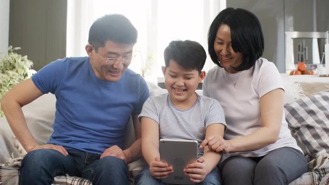 Tracking shot of Asian parents and school-age boy sitting on sofa and having fun while taking selfies on tablet