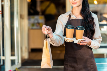 cropped image of waitress in apron holding two coffee in paper cups and paper bag near cafe