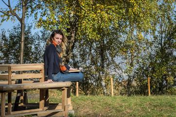 Young charming girl student sits on a bench in the autumn forest and reads a book