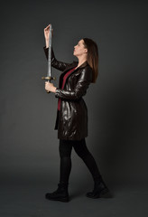full length portrait of brunette girl wearing long leather coat and boots. standing pose and holding a sword on grey studio background.