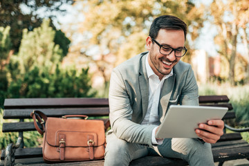 Smiling business man using tablet while sitting on the bench. - obrazy, fototapety, plakaty
