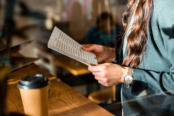 cropped view of businesswoman reading document at cafe with coffee to go