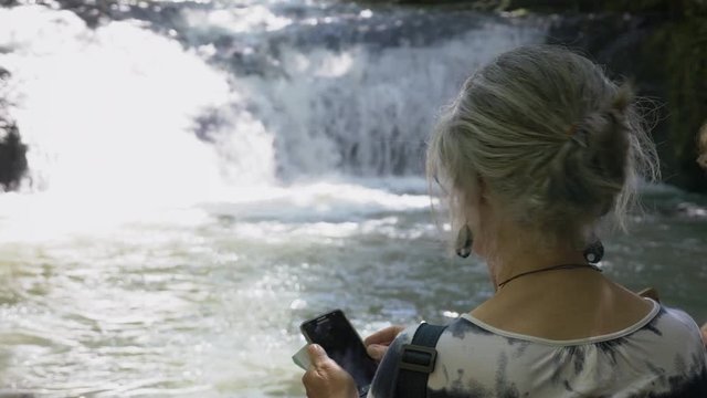Senior Woman Using Smartphone In Waterfall. Old lady using a smartphone enjoying the waterfall background.