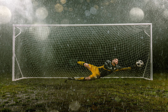 Dirty goalkeeper in flight catch the ball. Professional night rain stadium with football goal. Grass in the stadium wet from the rain