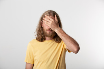 Young guy with a beard, mustache, blond hair down to the shoulders covers face with his hand. Isolated over white background