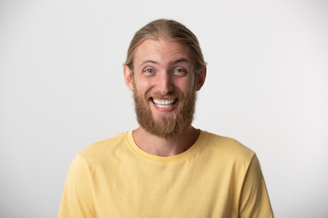 Obraz premium Cheerful young man with beard and blond hair put back smiles happily, has excited expression, dresssed casually, celebrates his anniversary or promotion at work, isolated over white studio background.