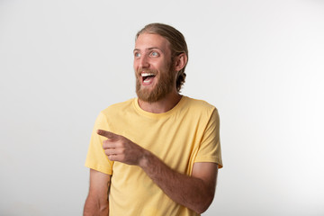 Cheerful young man with beard and blond hair put back with happy expression laughs, looks positively aside, points at blank copy space for your advertisement or promotional content.