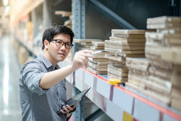 Young Asian man worker doing stocktaking of product in cardboard box on shelves in warehouse by using digital tablet and pen. Physical inventory count concept