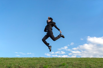 青空をバックにジャンプするスーツ姿の若いビジネスマン1人。元気・パワー・喜び・挑戦イメージ