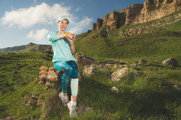 Portrait of a smiling young fitness girl in a cap and headphones checking her smart clock while sitting outdoors against a background of rocks. Looking at camera