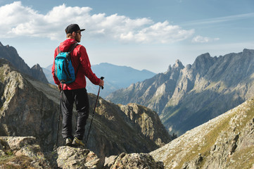 A bearded man in sunglasses and a cap with a backpack stands on top of a rock and looks into a rocky valley high in the mountains. The concept of tourism and easy trekking in the mountains outdoor © yanik88