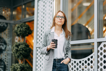 Girl blogger with coffee standing on the street, waiting for a meeting. Stylish and smiling girl. A happy buyer, a modern coffee shop.