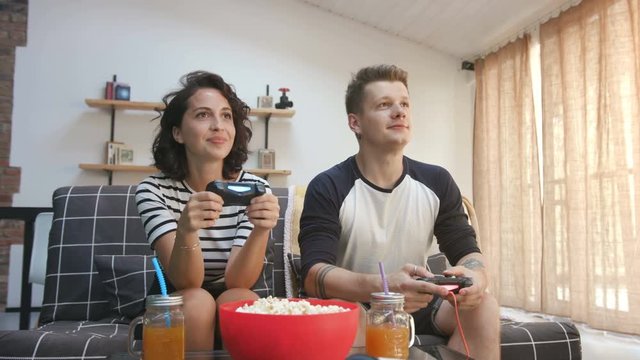 Good-looking Caucasian young woman sitting next to husband, playing video game. Popcorn and drinks on table near young couple. Indoors. Lifestyle.