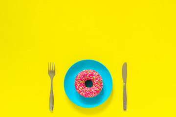Pink donut on blue plate and cutlery knife fork on yellow background.