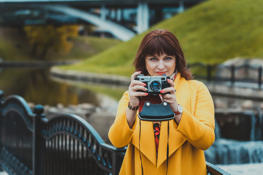 Woman with a camera. Adult forty year old beautiful woman in a yellow coat takes pictures in the city on the embankment by the river