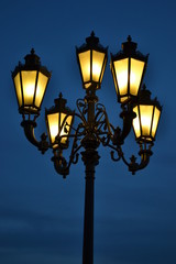 Fototapeta na wymiar Iron lamppost with five yellow lights at night on blue sky