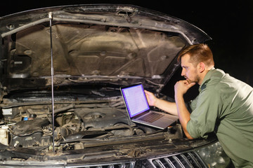 Mechanic in uniform looking intently at the laptop is connected to the motor vehicle for testing and adjustment. Mechanic carries out maintenance of the car. The removal of bugs of the engine.