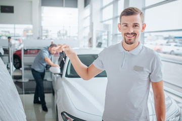 Obraz premium Picture of car deeler stands and looks on camera. He smiles and holds car key. Potential purchaser stands and leans on white car. He looks inside of it. with seriousness. They are in dealing store.