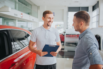 Picture of customer and seller stand together in front of beautiful red car. They look at each other and smile. Seller holds plastic tablet. They are on salon.