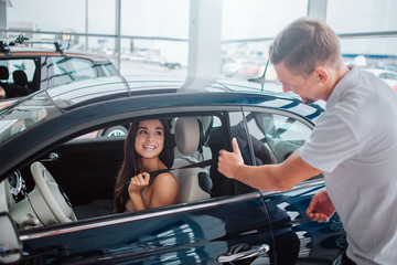Attractive and cheerful young woman sits in car. She looks at consultant and smile to him. Man holds door with one hand and smiles as well.