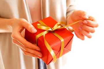 Young casual woman wearing pink shirt and long beige cardigan holding a beautiful present in shiny wrapping tied with golden bow. Giftwrapping concept. Background, copy space, close up.