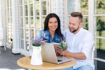 A happy married couple is resting in a cafe, laughing, enjoying delicious cocktails, using smartphones and a laptop. Modern family, people, technology, gadgets.