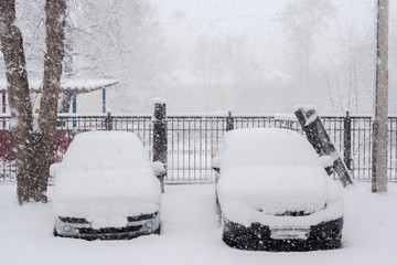 Two parked snow-covered cars in courtyard during plentiful snowfall.
