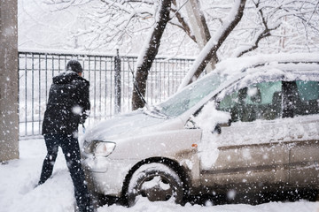 Man in dark clothes cleans his car during heavy snowfall. 