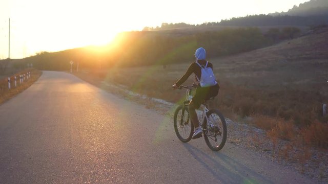 Muslim woman in hijab with backpack riding a bike on the road setting sun,rear view