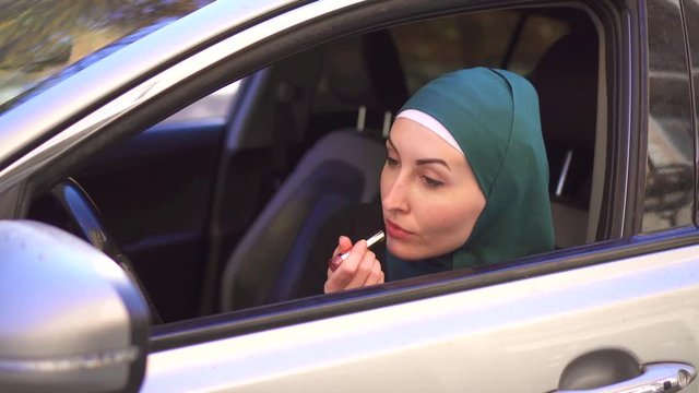 Muslim woman in a hijab sits in a car and paints her lips with lipstick