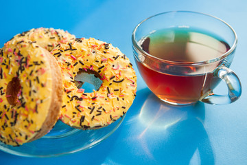 Black tea in glass cup and yellow sweets -  donuts biscuits on blue table with sunshine, lunch or breakfast