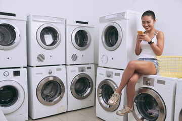 Pretty smiling Vietnamese woman sitting on washing machine in laundry room and texting friends