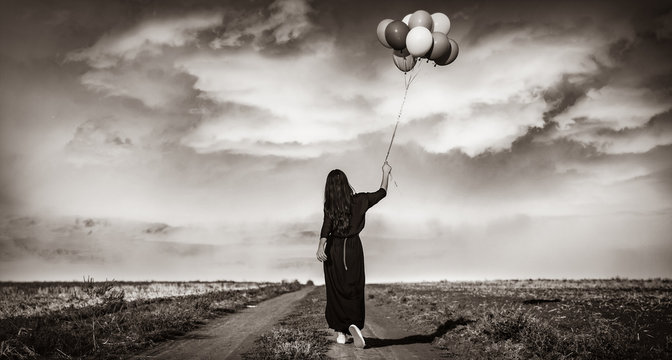 Beautiful girl in black dress with multicolored balloons on countryside . Image in black and white color