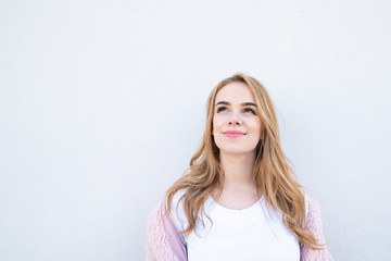 Fototapeta premium Attractive blond girl in a white T-shirt and a pink coat on a white background looks up. Сute girl on looks up on the background of a white wall. Copyspace.
