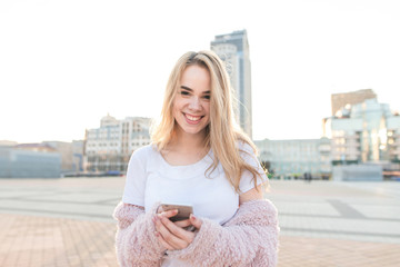 Happy, attractive young woman in a white T-shirt and a pink coat standing in the background of a city landscape with a smartphone in her hands, looking into the camera and smiling.