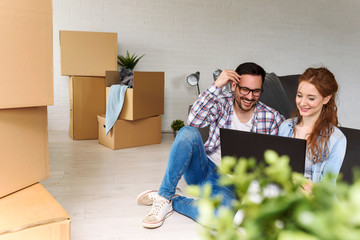 Beautiful young couple  shopping on-line using a laptop and smiling while sitting among cardboard boxes in their new apartment