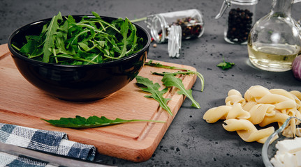 A view of food ingredients, rucola, garlic, dried tomatoes in brine, pasta, feta cheese. The concept of preparing dishes with pasta and rucola on a stone counter top. Food photography.
