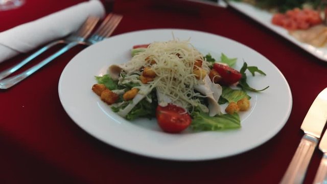 Slow motion of table in luxury restaurant preparing for dinner with best dishes. Close up of tasty classic salad cezar with tomato, chicken, mozzarella cheese on white rounded plate. Food concept.