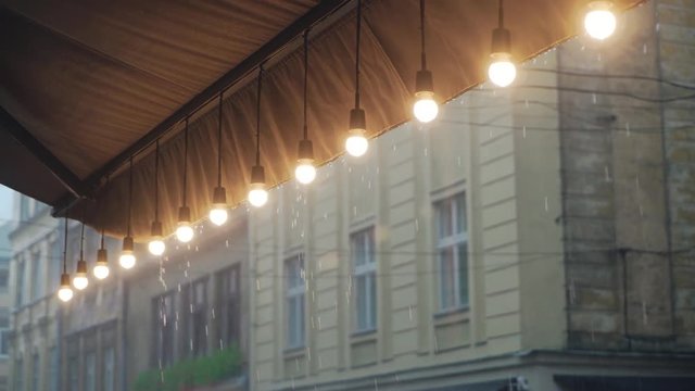 view on a row of burning light bulbs from the roof of a restaurant on the background of raindrops from the street. It's raining