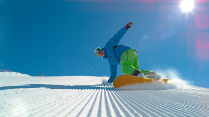 LOW ANGLE: Cheerful young male snowboarder carving a freshly groomed slope. - obrazy, fototapety, plakaty