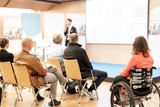 Rear view of nrecognizable woman on wheelchair participating in business conference talk. Business and entrepreneurship symposium. Speaker giving talk at business meeting. Audience in conference hall.