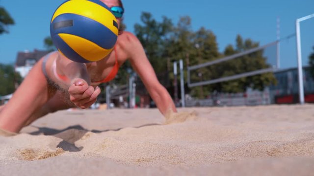 SLOW MOTION, CLOSE UP, DOF: Athletic Caucasian girl playing beach volleyball jumps into the sand and strikes the ball with her hand. Cinematic shot of fit female volleyball player saving a point.