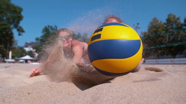 SLOW MOTION, CLOSE UP, DOF: Cheerful female teammates dive into the sand in effort to reach the ball during a beach volleyball tournament. Athletic women jump towards the ball and crash into the sand.