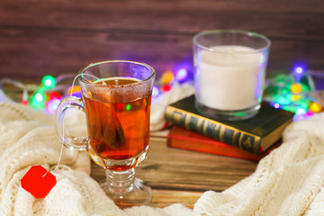Cozy winter still life: cup of hot tea and books, candle on wooden background and blurred garland lights