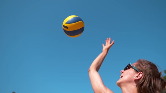 SLOW MOTION, CLOSE UP: Young Caucasian woman wearing sunglasses serves the ball in a fun beach volleyball game. Cinematic shot of happy female volleyball player striking the ball above her head.